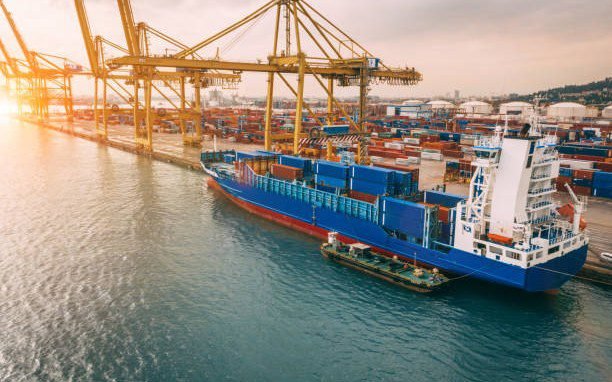 Container ship and port cranes seen from above — the sea freight leg to Fiji