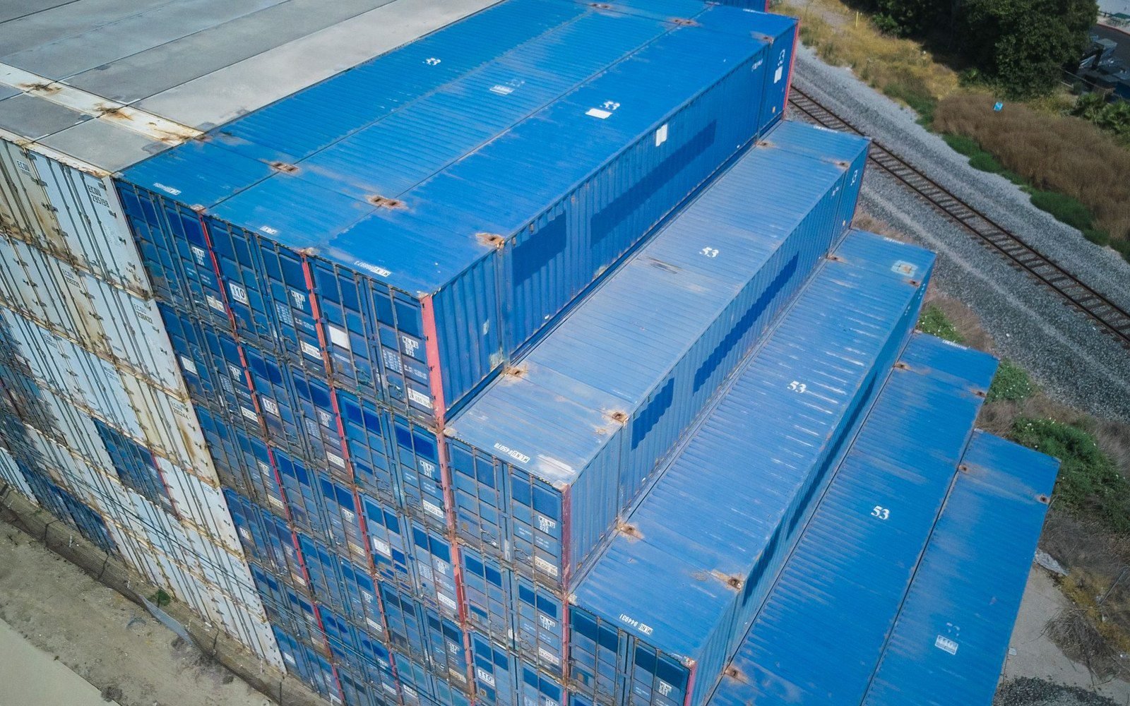 Aerial view of weathered blue shipping containers stacked beside a rail line (stock photo, Pexels 9716363)